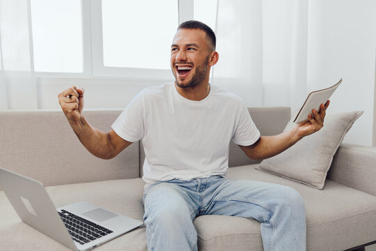 Happy man celebrating success at home with a laptop and notebook, showcasing enthusiasm and productivity in a cozy environment. Enjoying a vibrant and modern lifestyle.
