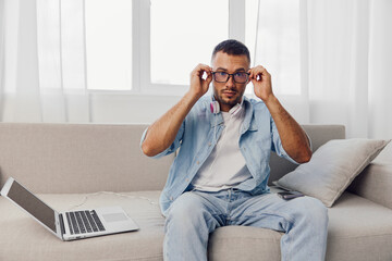 Focused man adjusting his glasses while sitting on a stylish sofa, surrounded by natural light....