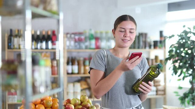 Girl in store scan QR code and study oil assortment. With help of mobile app, buyer receive information about olive oil product. She check manufacturer data in registry database of national agency