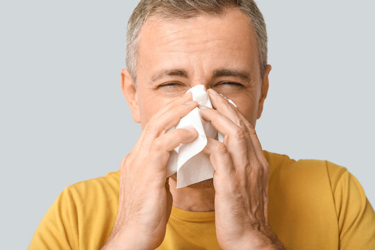 Ill mature man with tissue blowing nose on grey background