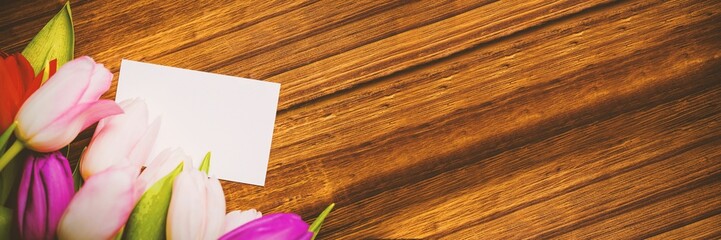Colorful tulip bouquet and blank card are resting on flat design warm wooden tabletop, copy space