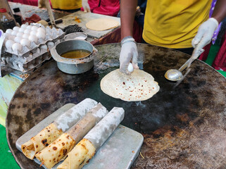 A vendor prepares a delicious egg roll by cracking a fresh egg onto a sizzling paratha on a hot iron tawa. Nearby, trays of neatly wrapped egg rolls, bursting with flavour, await serving.
