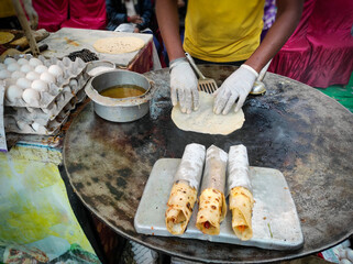 This image shows street food where a vendor prepares a delicious egg roll on a sizzling paratha on a hot iron tawa. Nearby, trays of neatly wrapped egg rolls, bursting with flavour, await serving.