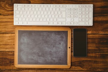 Flat wooden-framed blank chalkboard is lying on wood desk, with white keyboard and smartphone