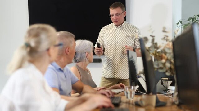 Middle-aged man teacher making explanation to elderly people sitting at computer table in IT training room