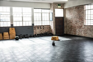 Single black kettlebell is sitting on rubber floor inside brick gym under grid windows, plyo boxes