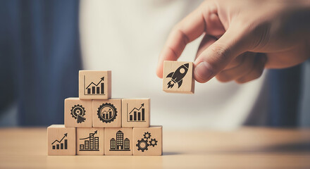 Close-Up of Hand Placing Final Wooden Cube with Rocket Icon to Complete a Stack of Business Symbols Representing Startup Growth

