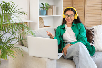 Beautiful young woman in headphones with laptop video chatting on sofa at home