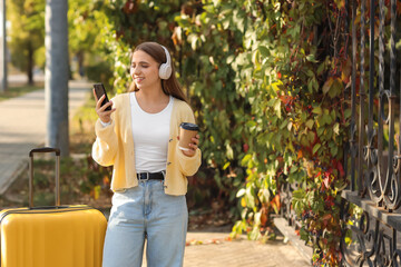 Female tourist with headphones and coffee using mobile phone in park on autumn day