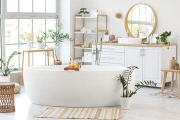 Interior of modern bathroom with bathtub, houseplants and cosmetic products