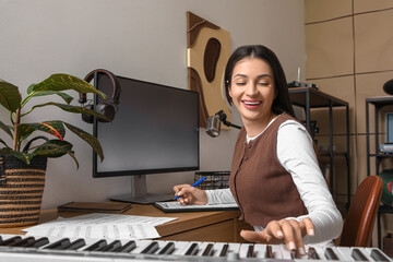 Beautiful young happy female composer writing notes while playing synthesizer in music studio