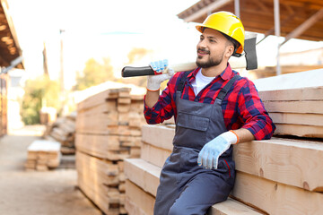 Young carpenter in hardhat with axe near wooden planks at sawmill