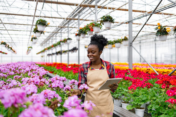 An African-American woman tends to flowers in a greenhouse, using a tablet to monitor growth and development.
