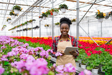 A smiling African woman in a greenhouse with a tablet, surrounded by vibrant flowers and plants, and other potted plants.