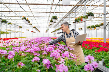 A man in a greenhouse examines flowers, holding a laptop, surrounded by vibrant blooms.