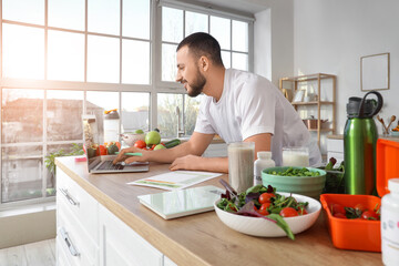 Sporty young man with fresh vegetables, laptop and meal plan in kitchen. Healthy food concept