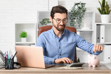 Young businessman with calculator putting coin into piggy bank at table in office