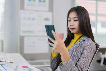 Young Asian businesswoman drinking coffee and using smartphone in modern office with charts and...