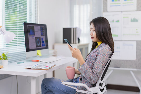 Young Asian businesswoman using smartphone and drinking coffee while sitting at her desk in modern office, working with charts and graphs showing on computer screen