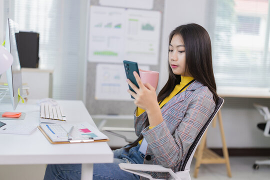 Young Asian businesswoman drinking coffee and using mobile phone while sitting at desk in modern office, working with financial report and computer