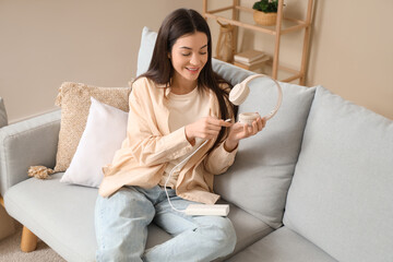 Beautiful young woman with modern power bank and headphones sitting on sofa at home