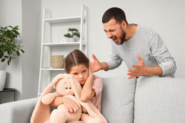 Scared girl with toy sitting on sofa and her angry father at home. Domestic violence concept