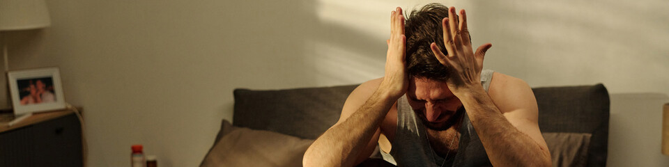 Caucasian young adult man sitting on bed holding head in hands appearing distressed, struggling...