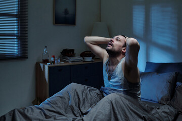 Caucasian young adult man holding head with both hands appearing distressed in dimly lit bedroom...