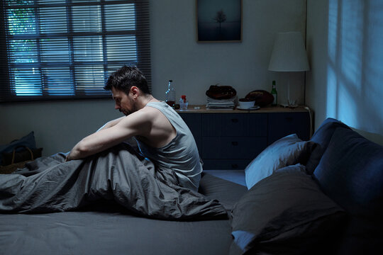 Caucasian young adult man sitting on bed holding knees appearing distressed in dimly lit bedroom, facing away from camera, conveying emotional struggle related to family problems