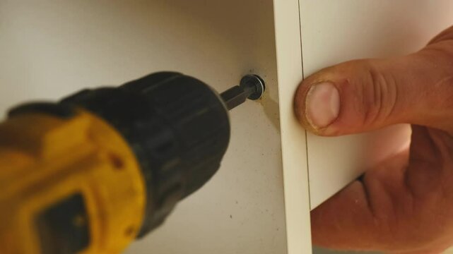 Worker tightens screw to fix cabinet panel in place. Installer adjusts speed of screwdriver and tightens screw to attach board of cabinet