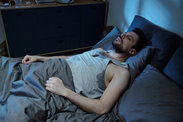 Caucasian young adult man lying in bed sweating heavily during sleep, appearing distressed and...