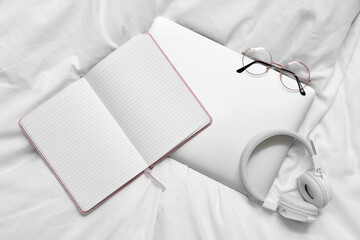 Laptop with eyeglasses, headphones and blank notebook on bed, top view