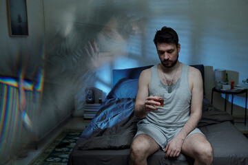 Caucasian man sitting on bed holding glass of alcohol looking down appearing distressed with...