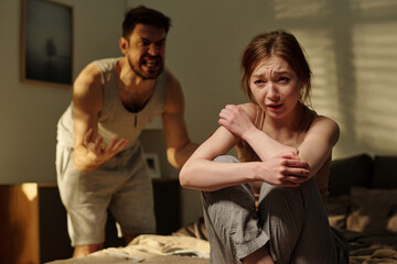 Man shouting while standing behindwoman sitting on bed hugging knees looking distressed,...