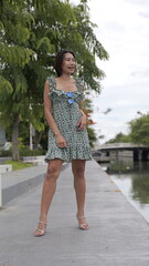 Asian woman stands in a floral dress and stylish high-heeled sandals by a serene waterway,...