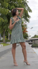 Asian woman holding her sunglasses stands in a floral dress and stylish high-heeled sandals,...