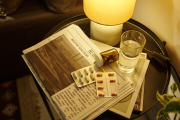 Close-up showing glass of water, pill bottle, blister packs of medication and stack of newspapers...