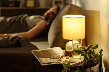 Caucasian man lying in bed appearing unwell, bedside table holding glass of water, blister packs of...