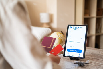 Woman is booking a flight online using a travel app on a tablet, holding her passport and credit card, ready to embark on a new adventure, with a small globe and airplane model nearby