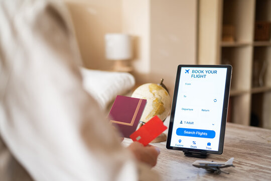 Woman is booking a flight online using a travel app on a tablet, holding her passport and credit card, ready to embark on a new adventure, with a small globe and airplane model nearby - Powered by Adobe