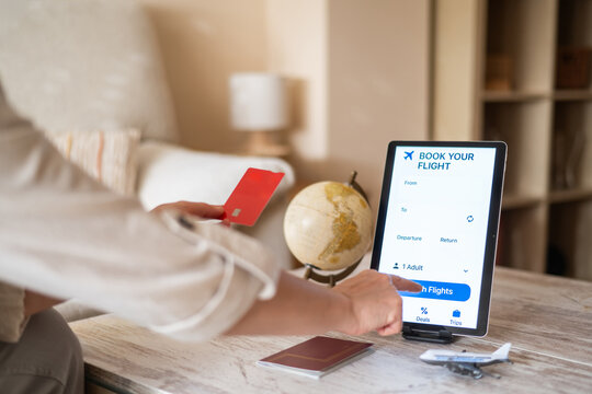 Woman holding credit card and using tablet for booking flight tickets online at home, with passport and airplane model placed on table, showcasing online travel booking and digital tourism