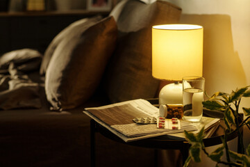 Nightstand holding glass of water, several blister packs of pills, newspapers and lamp beside bed,...