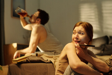 Woman sitting on floor holding phone with anxious expression while young man sitting on bed drinking alcohol from bottle in background suggesting family conflict