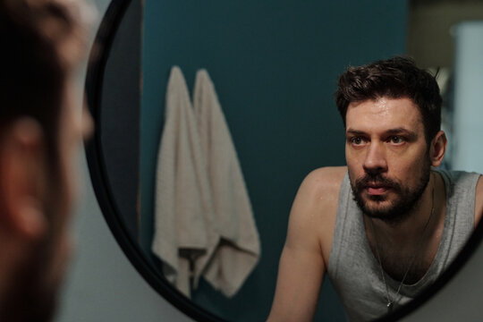 Caucasian young adult man staring into mirror with serious expression, leaning forward with hands on sink, appearing deep in thought in bathroom setting