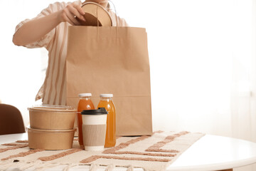Young woman taking container from paper bag on table in kitchen. Food delivery service