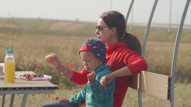 A woman sits at a table, reaching for food, with a young child resting against her side. They are enjoying a tranquil outdoor meal by a field, peaceful interlude during their travels in Crimea.