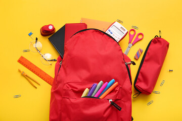 Red school backpack with different stationery supplies and eyeglasses on yellow background