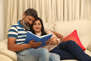 Indian couple man and women on cozy couch sofa and relaxing in her living room reading book in Lifestyle Concept. Husband wife spend time with each other and having fun