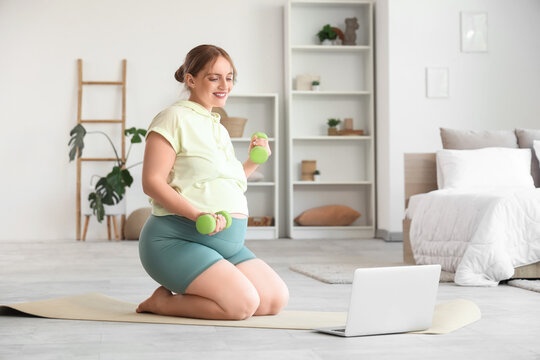 Sporty pregnant woman training with dumbbells and laptop in bedroom