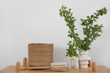 Vases of green blossoming branches with basket and massage brush on table near white wall in...